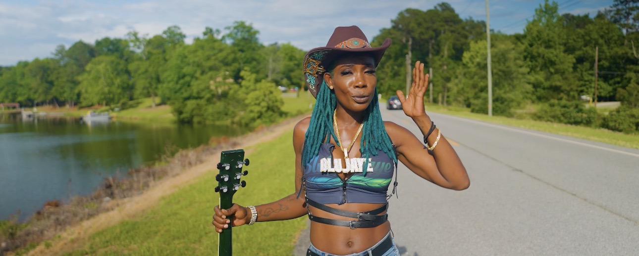 Woman with long braids wearing a hat and striped top holding a guitar by a lake, on a road with trees in background.
